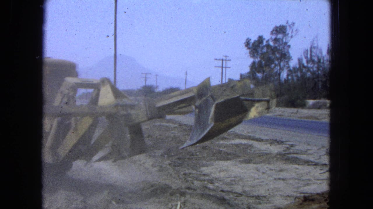 Yellow bulldozer blade pushing dirt beside a rural road with poles and trees.