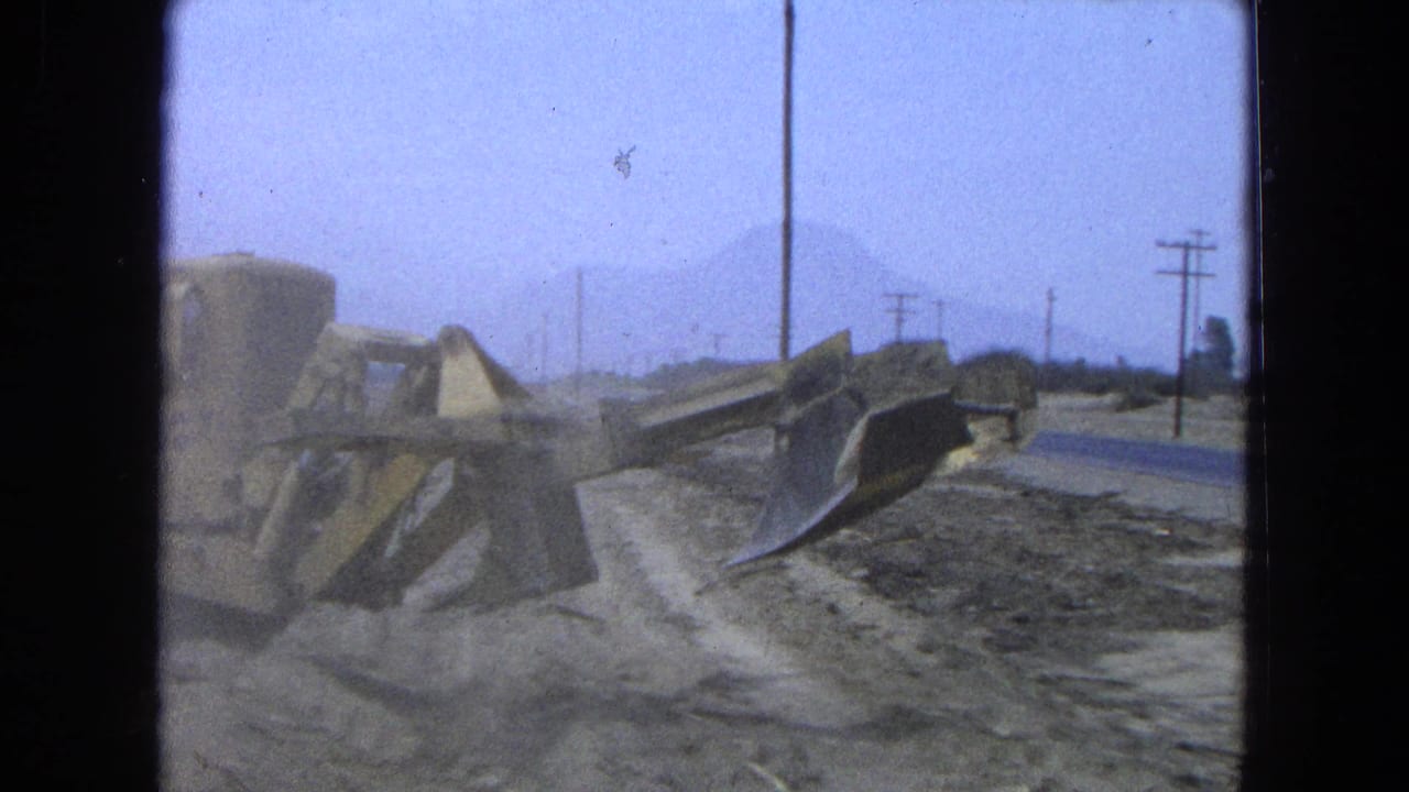 1965 Pueblo, Mexico frame with a yellow bulldozer working on a dirt road shoulder, utility poles, haze, and mountains in the distance.