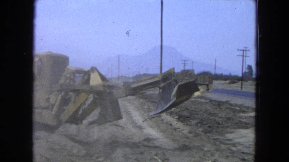 1965 Pueblo, Mexico frame with a yellow bulldozer working on a dirt road shoulder, utility poles, haze, and mountains in the distance.
