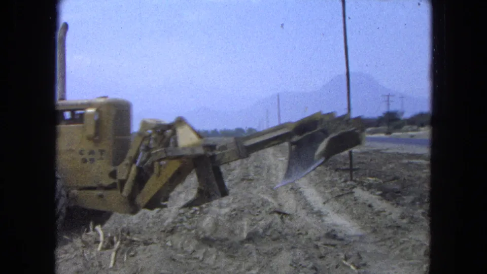 1965 Pueblo, Mexico home-movie frame showing the side of a yellow bulldozer and blade beside a rough dirt path with mountains beyond.