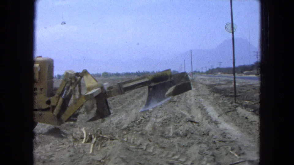 1965 Pueblo, Mexico frame showing a bulldozer blade over a mound of dirt near roadside poles, with a hazy mountain ridge in the background.