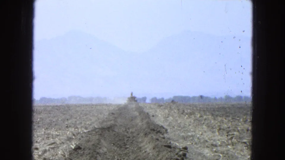 1965 Pueblo, Mexico 8mm frame with a dusty trench crossing open farmland and a distant excavator set against a hazy mountain backdrop.