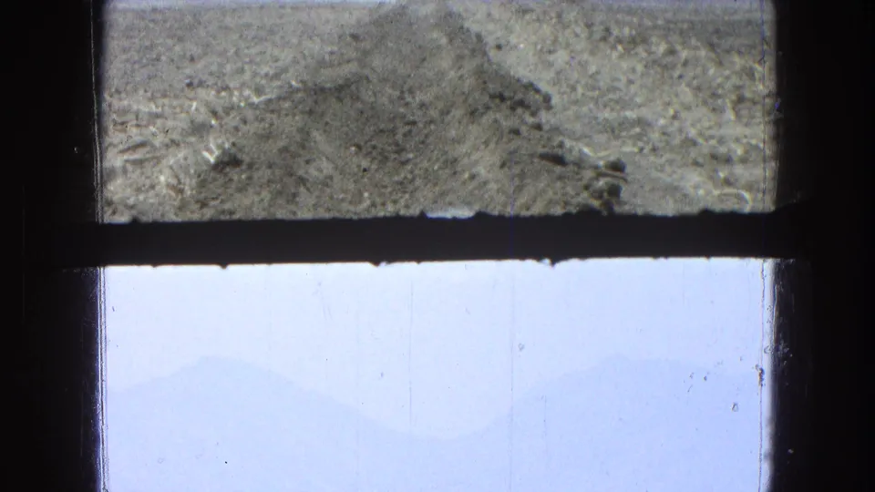 1965 8mm Pueblo, Mexico frame with rough earth above a dark divider and pale blue sky with faint mountain silhouettes below.