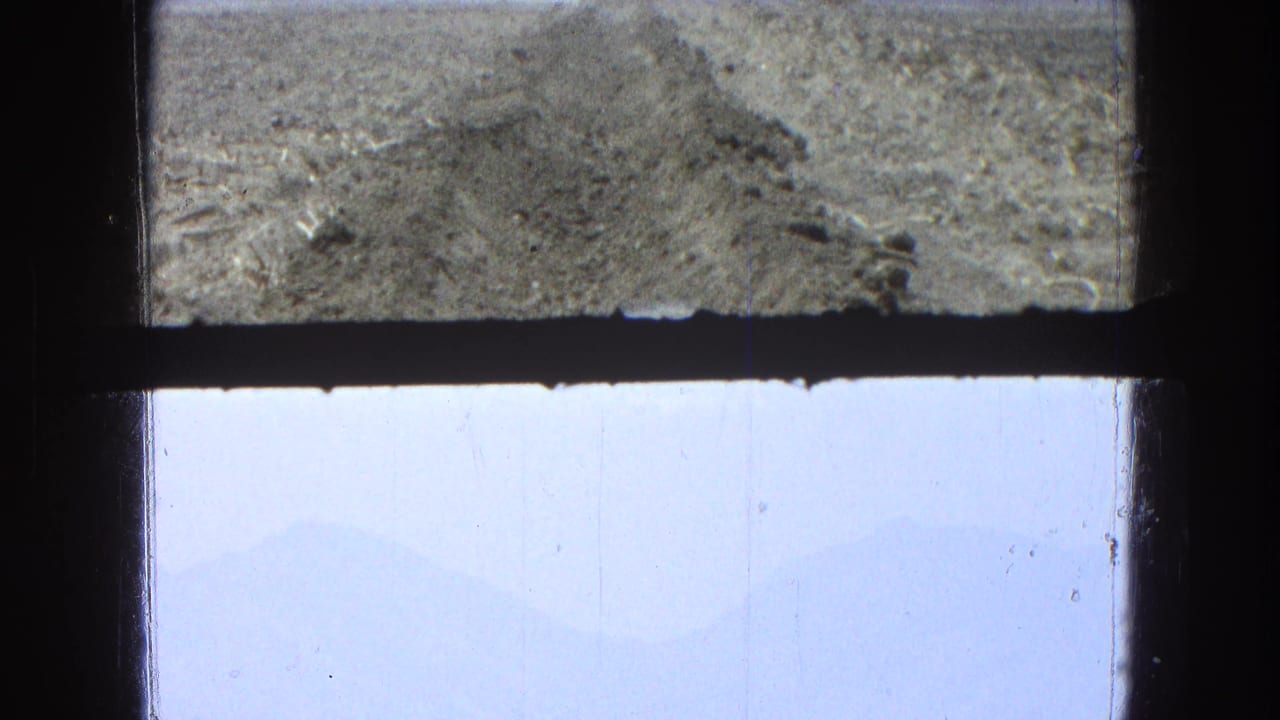1965 8mm Pueblo, Mexico view divided by a dark frame, showing textured ground, bright sky, and soft distant mountain forms.