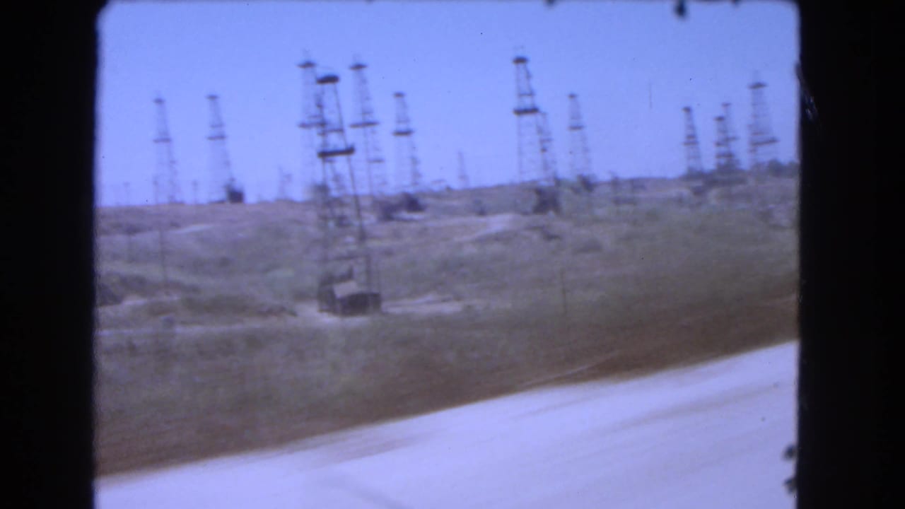 Roadside view of oil derricks on a dusty hillside under a blue sky.