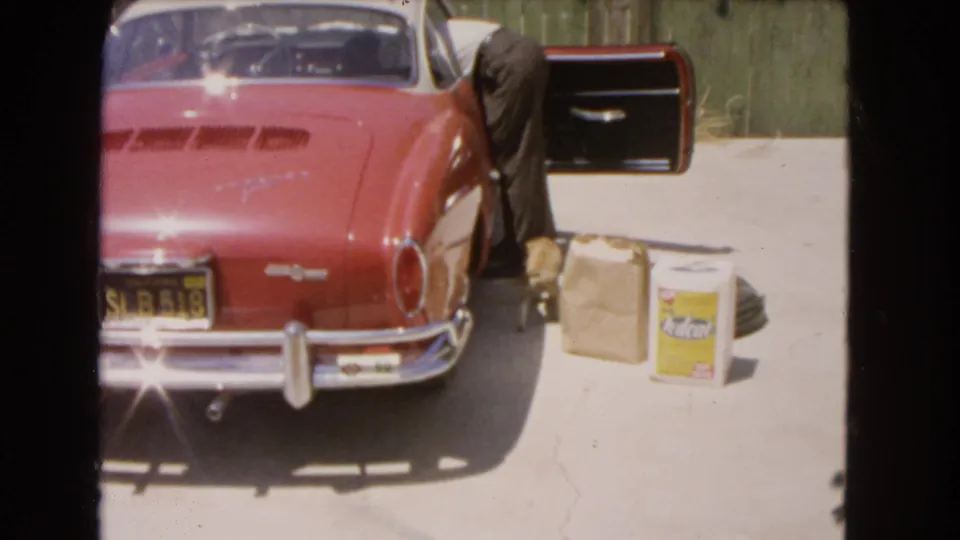 An adult in dark pants leans into the open door of a red Volkswagen Karmann Ghia while a small dog stands by the rear bumper.