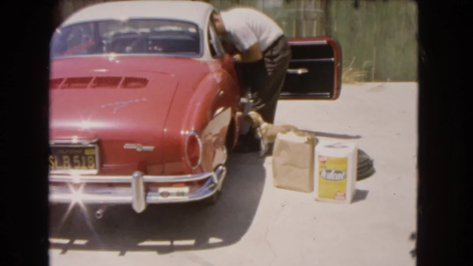 Adult in a white shirt and dark pants bends near the open door of a red Volkswagen Karmann Ghia as the small dog stands close on the driveway.