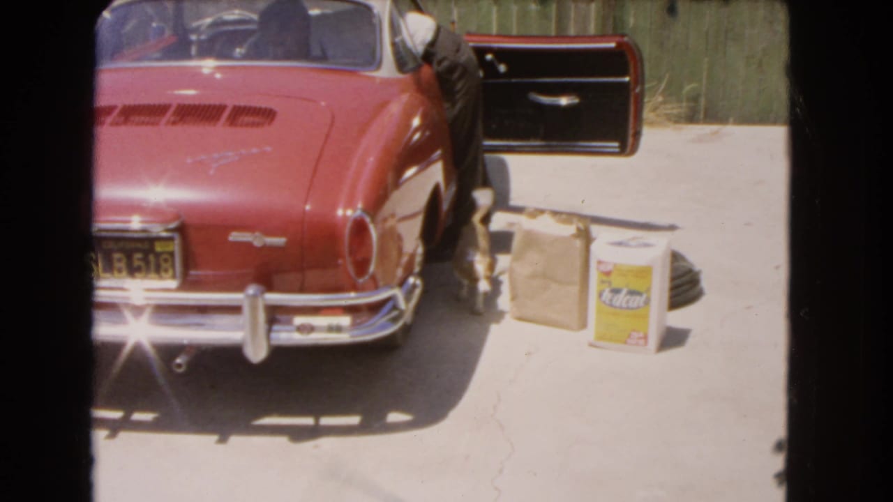 1966 Barstow 8mm frame of a red Volkswagen Karmann Ghia with an open door, chrome bumper, paper bag, and yellow container in a driveway.