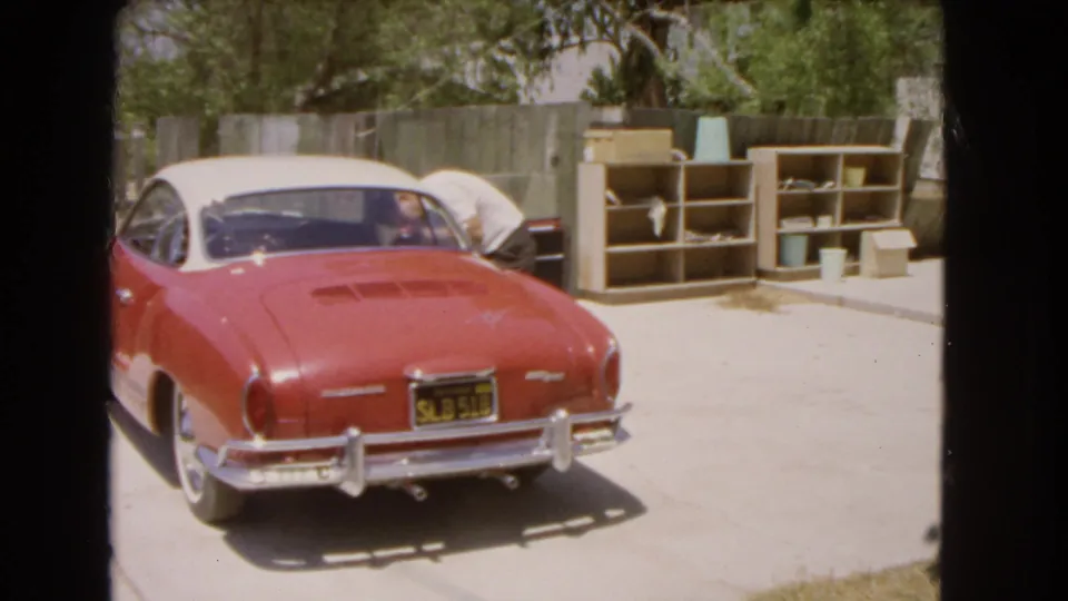 1966 Barstow frame of a person loading or arranging items in a red Karmann Ghia beside outdoor shelves, boxes, and a fenced driveway.