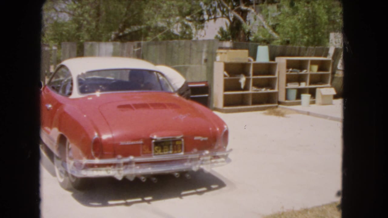 Red and white classic coupe parked in a sunny driveway beside shelves and a wooden fence.