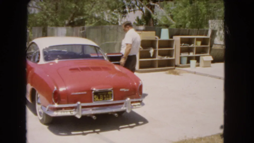 In 1966 Barstow footage, a man stands near the driver's door of a red and white Volkswagen Karmann Ghia in a backyard driveway.