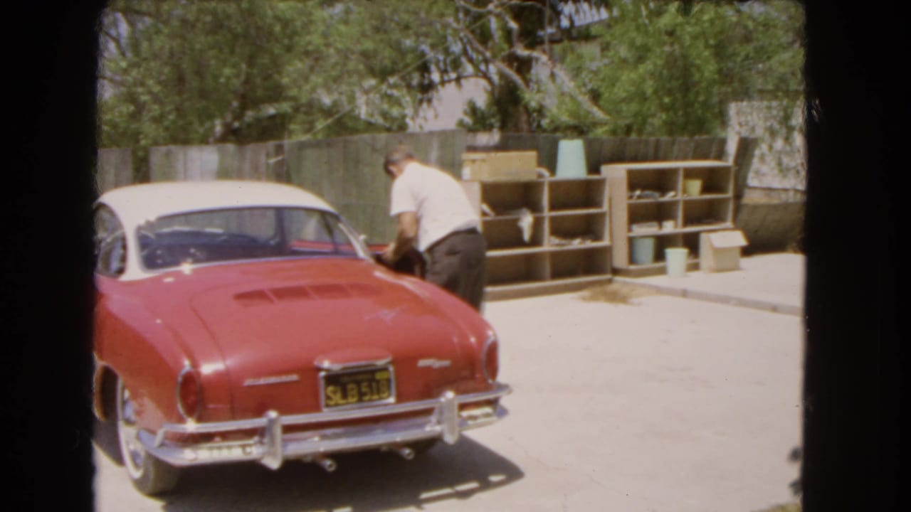 Man beside a red and white vintage car in a sunny driveway.