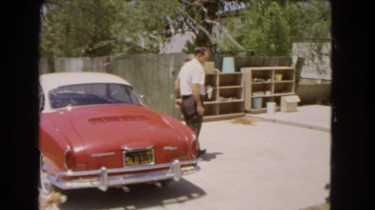 The man walks away from the red and white Volkswagen Karmann Ghia, with outdoor shelving and a fence behind him.