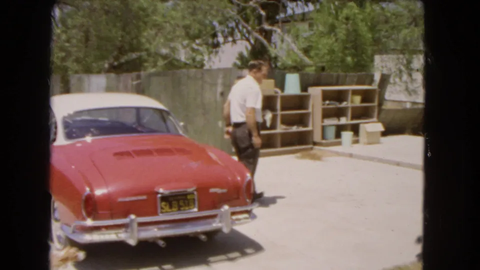 The man walks away from the red and white Volkswagen Karmann Ghia, with outdoor shelving and a fence behind him.