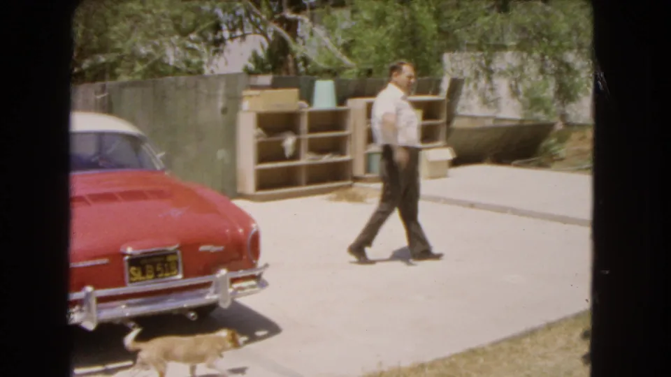 A man crosses the sunlit driveway while a small dog appears near the rear of the vintage car.