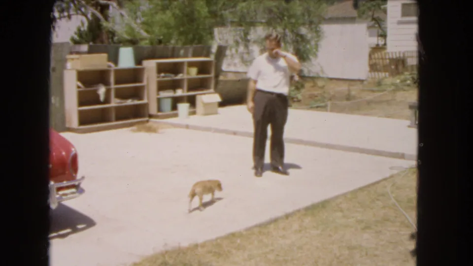 1966 Barstow home-movie frame showing an adult man on a sunny backyard patio as a small dog walks nearby.