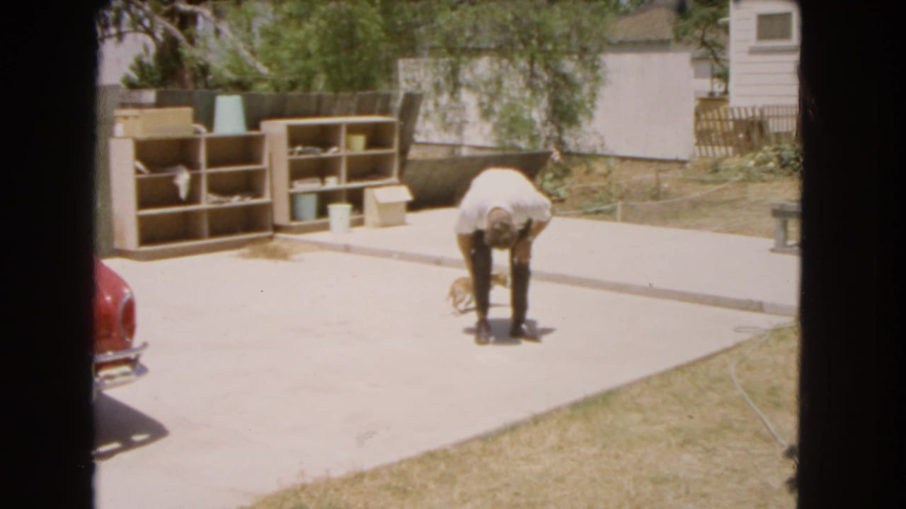 1966 Barstow backyard frame with the man bending down toward the small dog in bright sun.