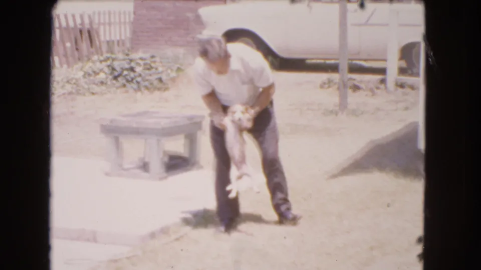 1966 Barstow backyard frame showing the man holding the small dog upright during a casual pet interaction.