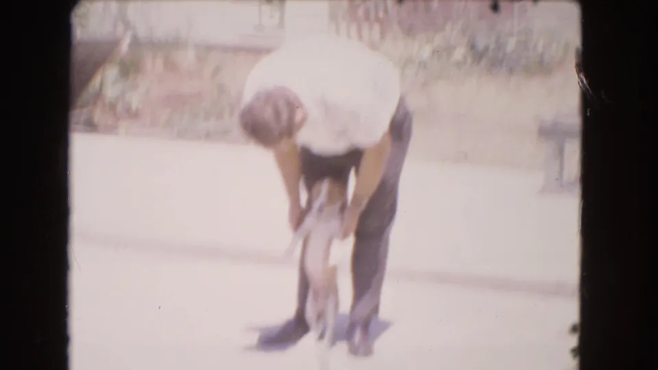 In 1966 Barstow home-movie footage, a man in a white shirt bends over a small puppy on a sunlit driveway.