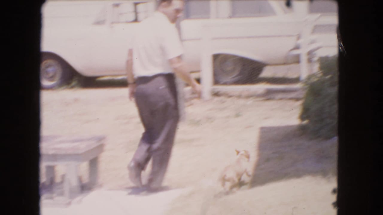 1966 Barstow home-movie frame showing an adult man in a white shirt crossing a sunlit yard with a small light-colored dog nearby.