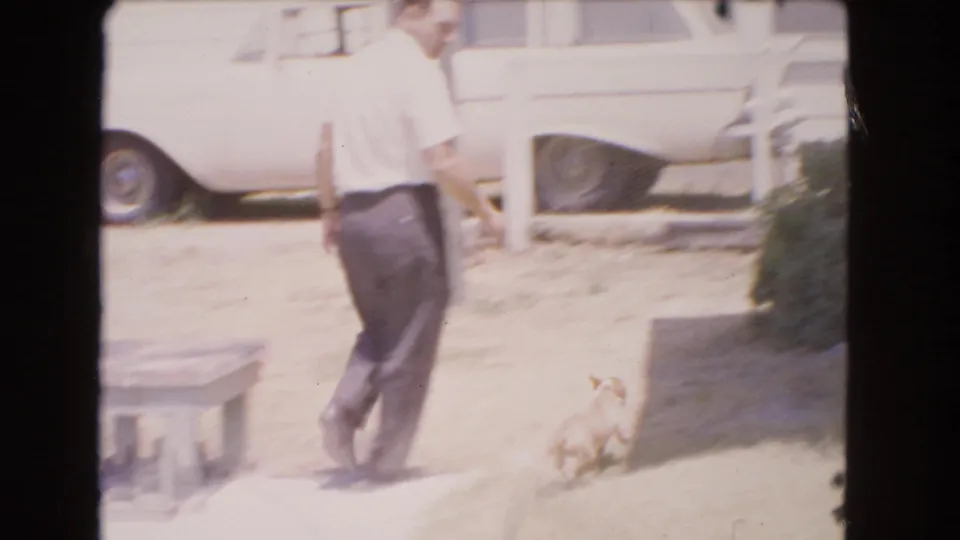 1966 Barstow home-movie frame showing an adult man in a white shirt crossing a sunlit yard with a small light-colored dog nearby.