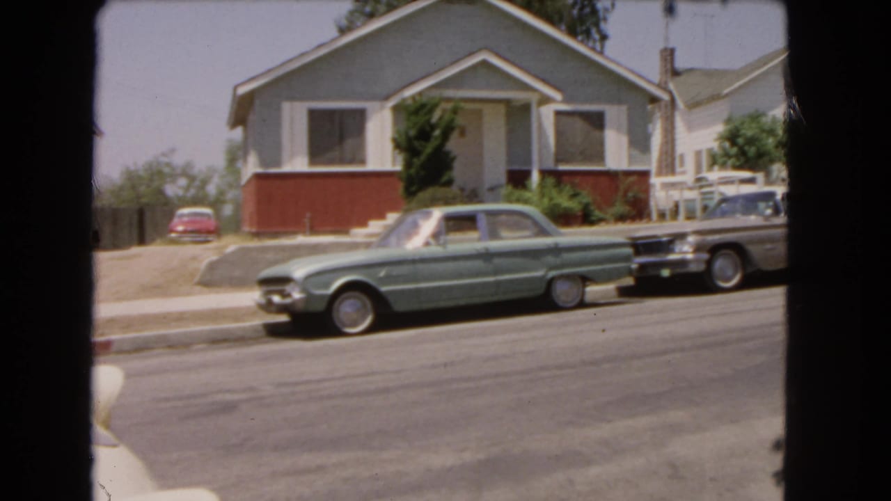 1966 Barstow home-movie frame showing a green sedan and darker car parked outside a light-sided house on a sunny street.