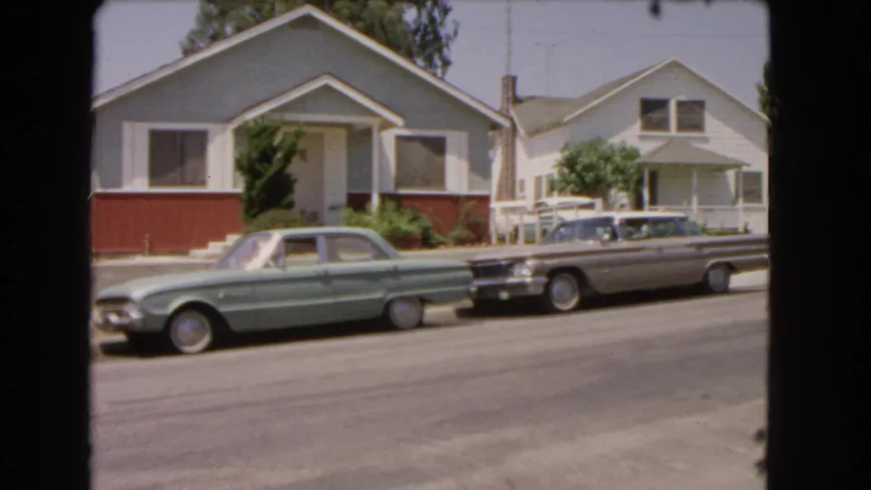 1966 Barstow home-movie still showing parked classic cars, a light blue house, and a neighboring home across a sunny street.