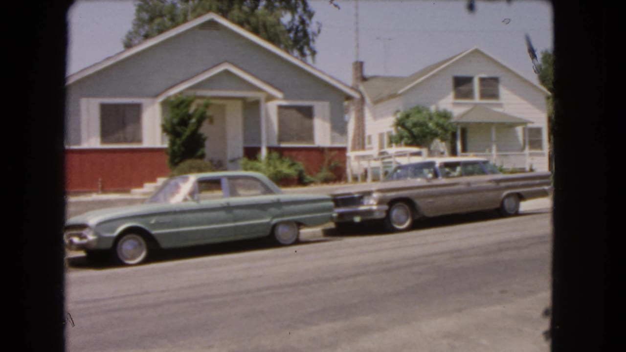Two parked cars on a sunny residential street in front of single-story houses.
