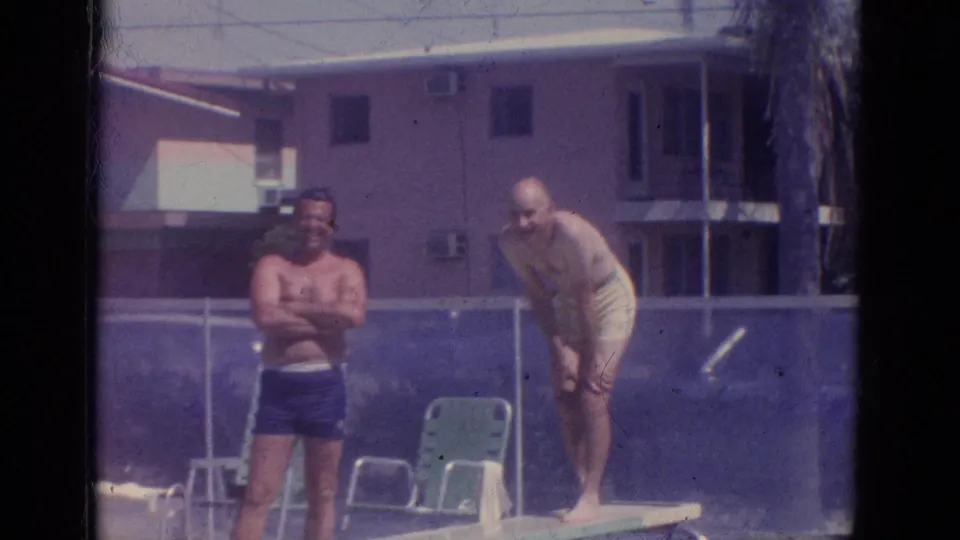 1966 Florida home-movie frame with two adult men at a resort pool diving board, captured in soft faded color.