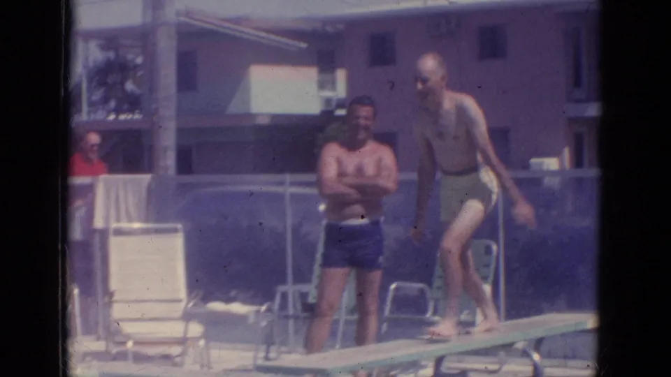 1966 Florida resort pool frame showing two adult men at the diving board with lounge chairs and pool fencing behind them.
