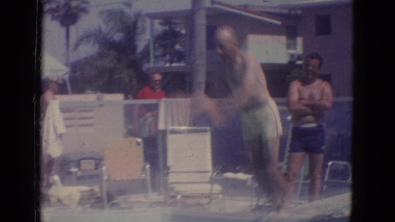 Adult man diving into a pool while people watch near lounge chairs.