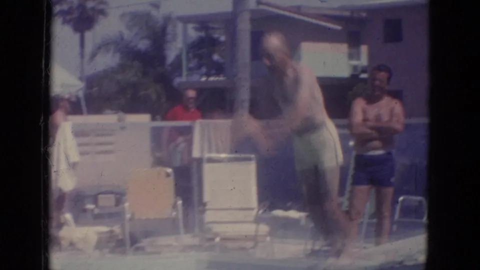 1966 Florida 8mm frame of an adult man jumping from a diving board as poolside spectators and chairs sit behind him.