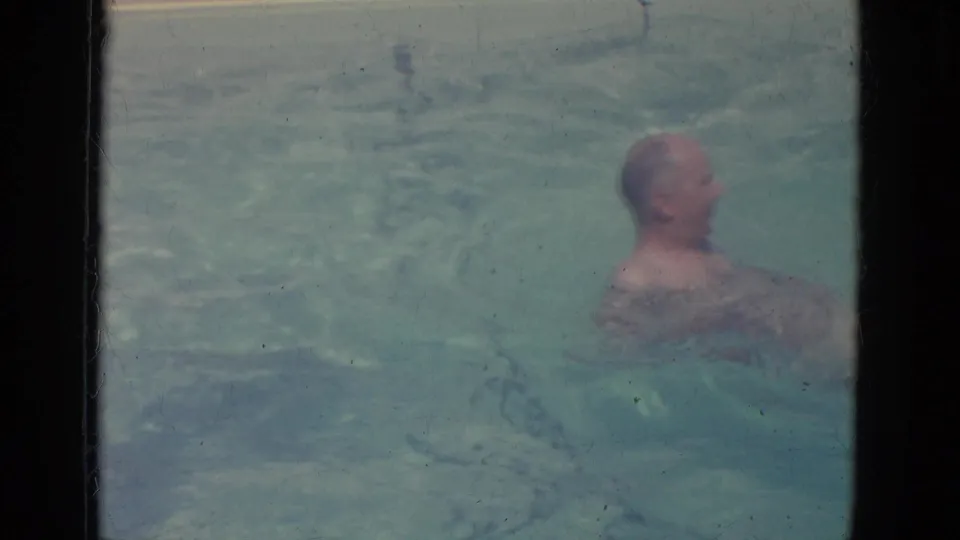 1966 Florida home-movie frame of a bald adult man resurfacing and swimming in clear pool water.