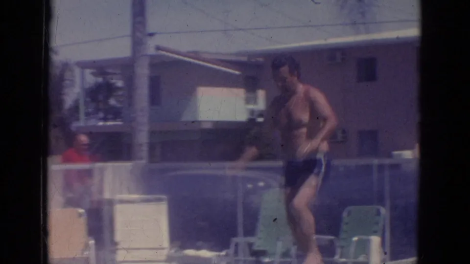 1966 Florida 8mm frame of an adult man in dark swim trunks preparing near the diving board with poolside chairs behind him.