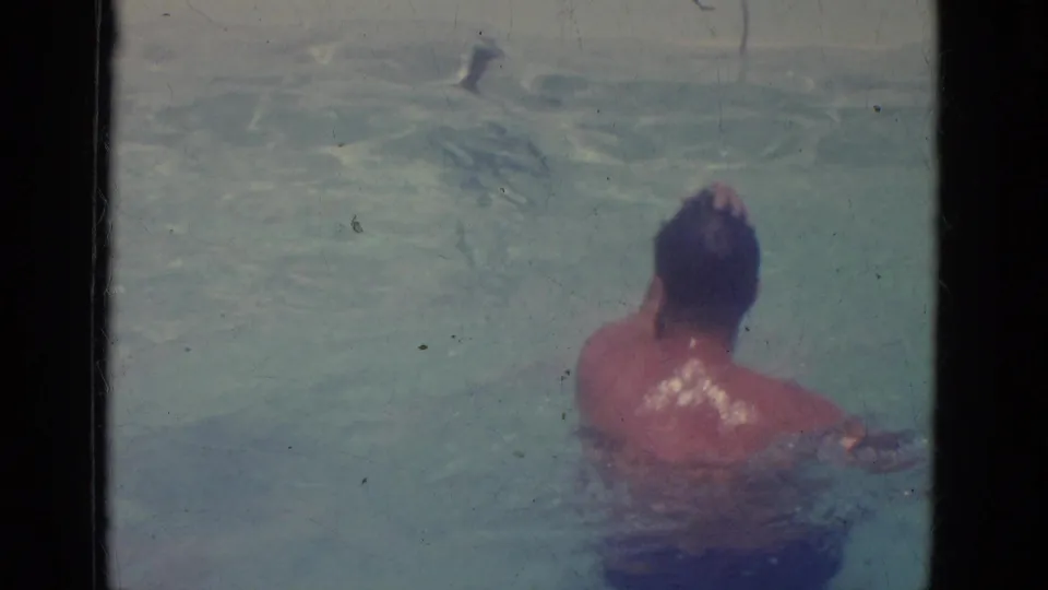 An adult man resurfaces in the outdoor pool, viewed from behind in soft 1966 Florida home-movie footage.