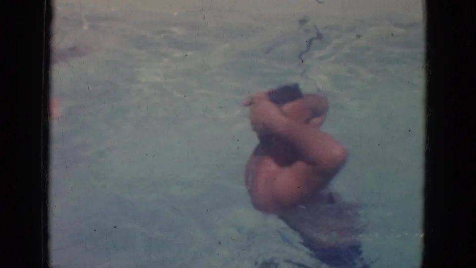 An adult man pauses in the pool with both hands on his head, filmed in a soft, faded 8mm home-movie frame.
