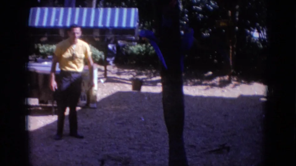 A 1966 8mm view at Busch Gardens shows a man in a yellow shirt standing near a striped awning, trees, and bird display stands.