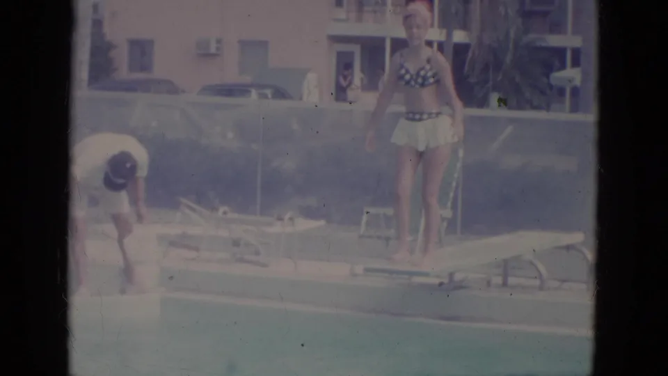 1966 Florida 8mm poolside frame with a woman on a diving board, another person nearby, lounge chairs, parked cars, and a building behind.