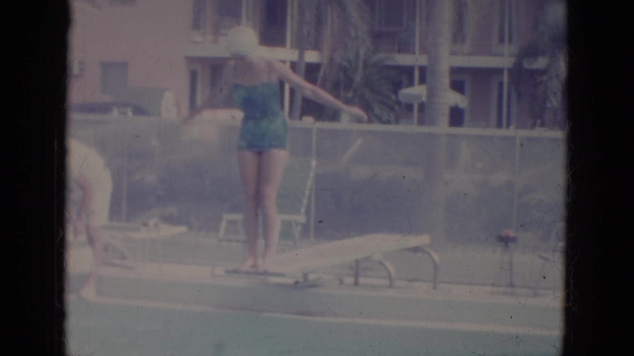 Woman in a teal swimsuit stands at a pool edge with arms out beside lounge furniture and buildings.