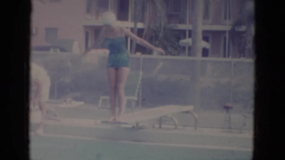 In 1966 Florida home-movie footage, a woman in a teal swimsuit and light swim cap prepares to dive beside a pool with chairs and buildings behind her.
