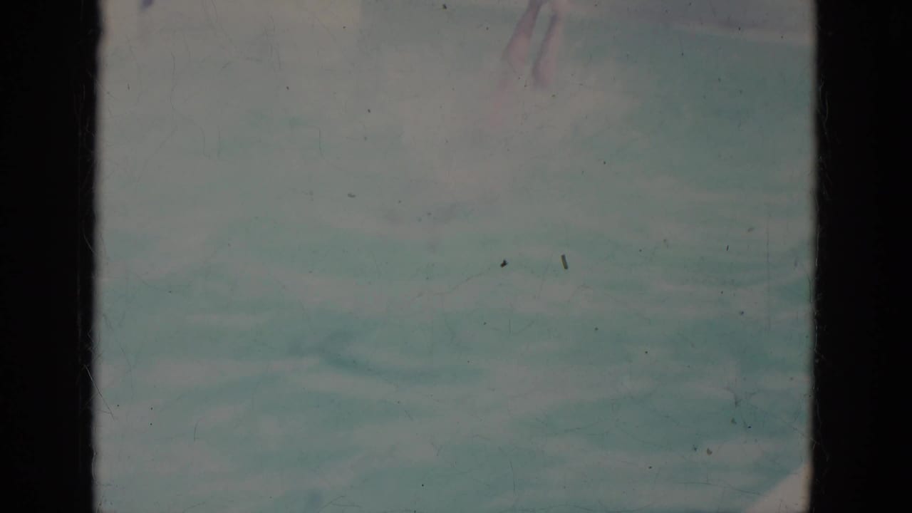 Faded 8mm frame shows blue-green pool water just after the dive, with part of the swimmer barely visible near the top of the frame.