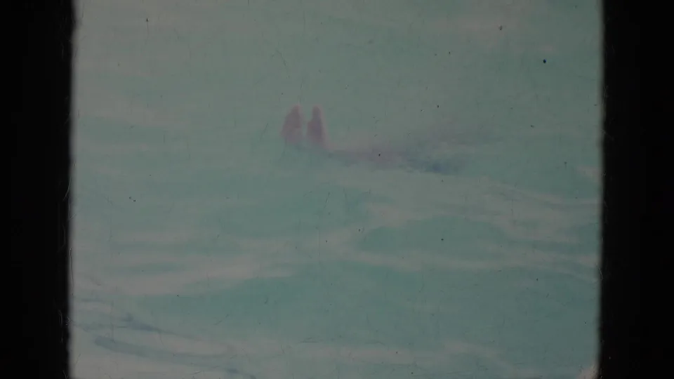 Underwater 8mm view of a swimmer moving through a blue-green pool, with the body softened by water, fading, and film grain.