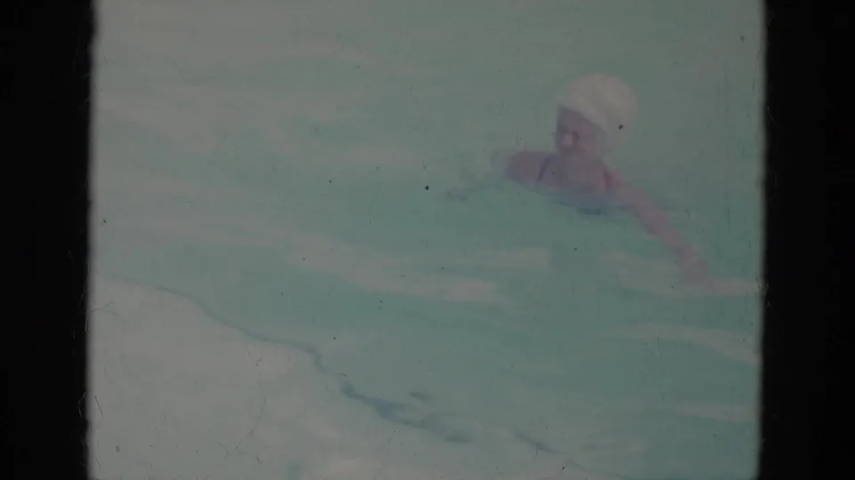 A swimmer wearing a light swim cap surfaces in the pool during 1966 Florida home-movie footage, with facial detail softened by water and film fade.
