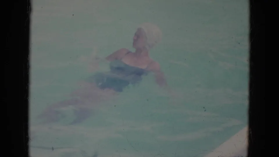 1966 Florida home-movie frame showing a woman in a swim cap and dark swimsuit floating in a turquoise pool.