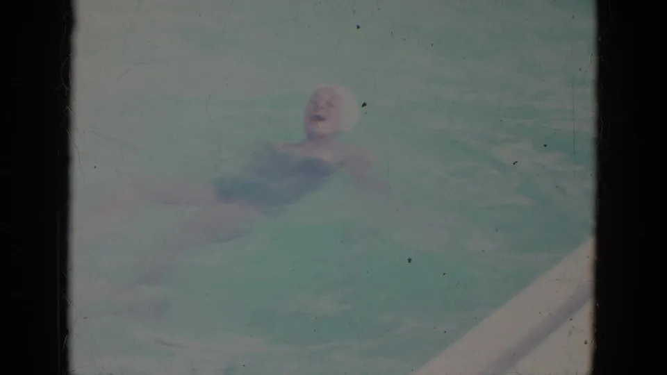 Super 8 frame from the 1966 pool scene showing a woman in a white cap facing the camera while swimming.