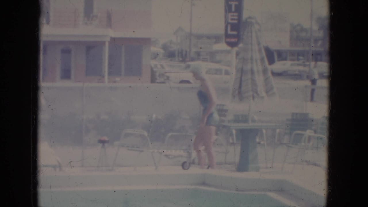 Woman in swimsuit and swim cap walks beside a motel pool toward a diving board.