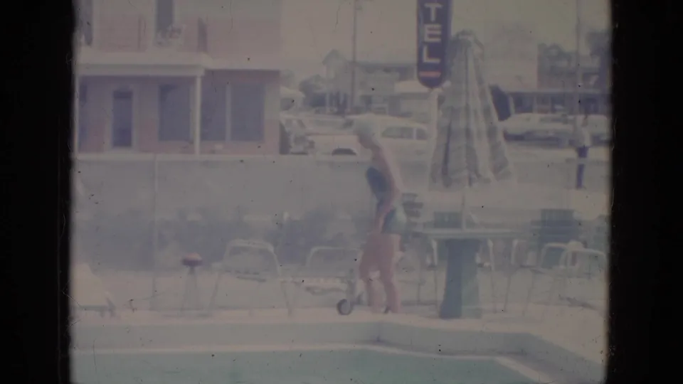 1966 8mm frame of a woman in a one-piece swimsuit and swim cap near a motel pool, with chairs, umbrella, parked cars, and hotel sign behind her.