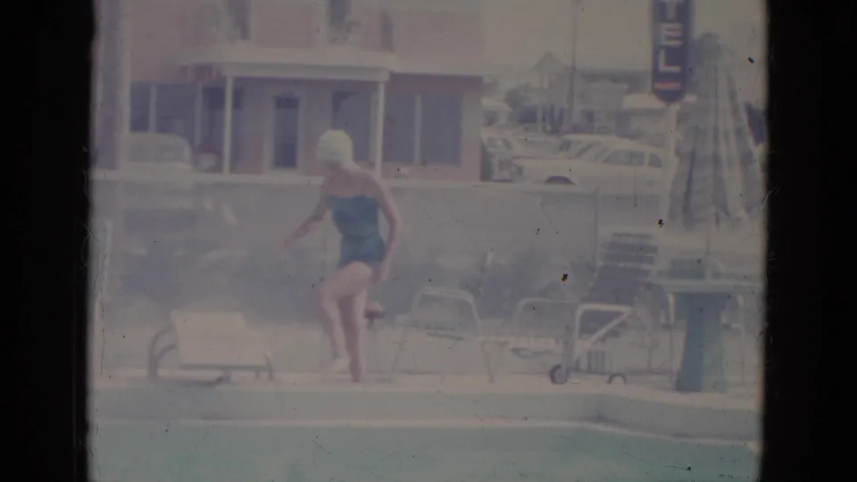 1966 Florida home-movie frame showing a woman in a swimsuit and cap approaching a low diving board at a motel-style pool.