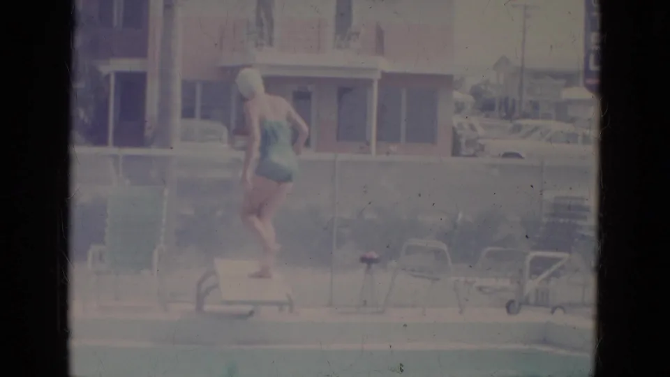 Faded 8mm frame of a woman on a low diving board at a 1960s Florida pool, with chairs, umbrella, and motel building behind her.