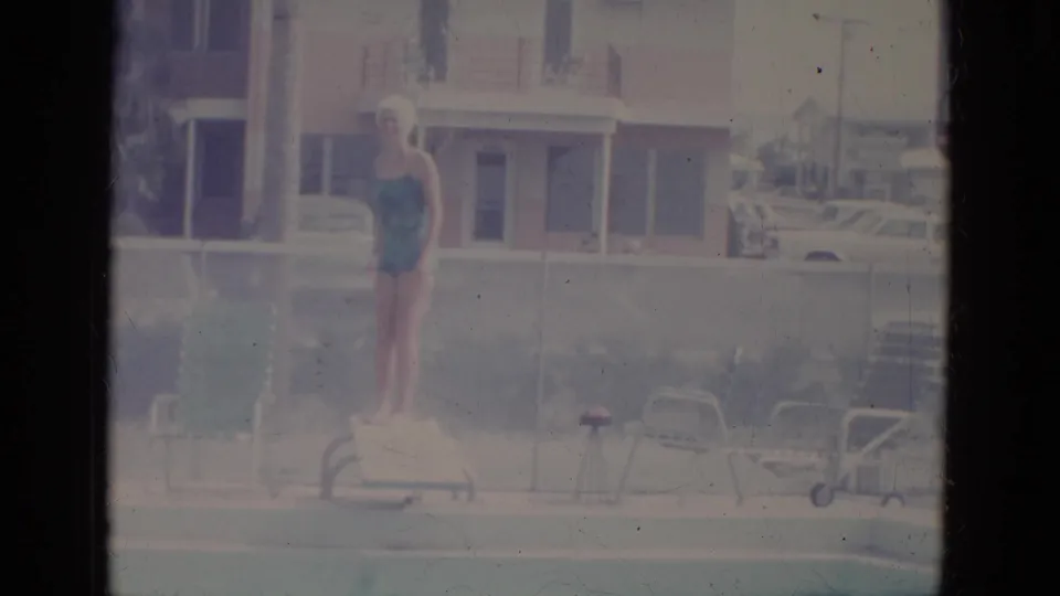 1966 home-movie frame of a woman in a swimsuit and swim cap standing on the diving board at a motel pool in Florida.